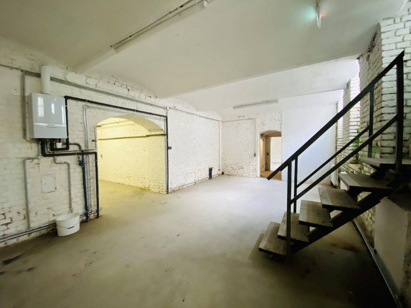 Kellerraum mit freiliegenden Rohren, Heizungsanlage, Betonboden und einer Metalltreppe.