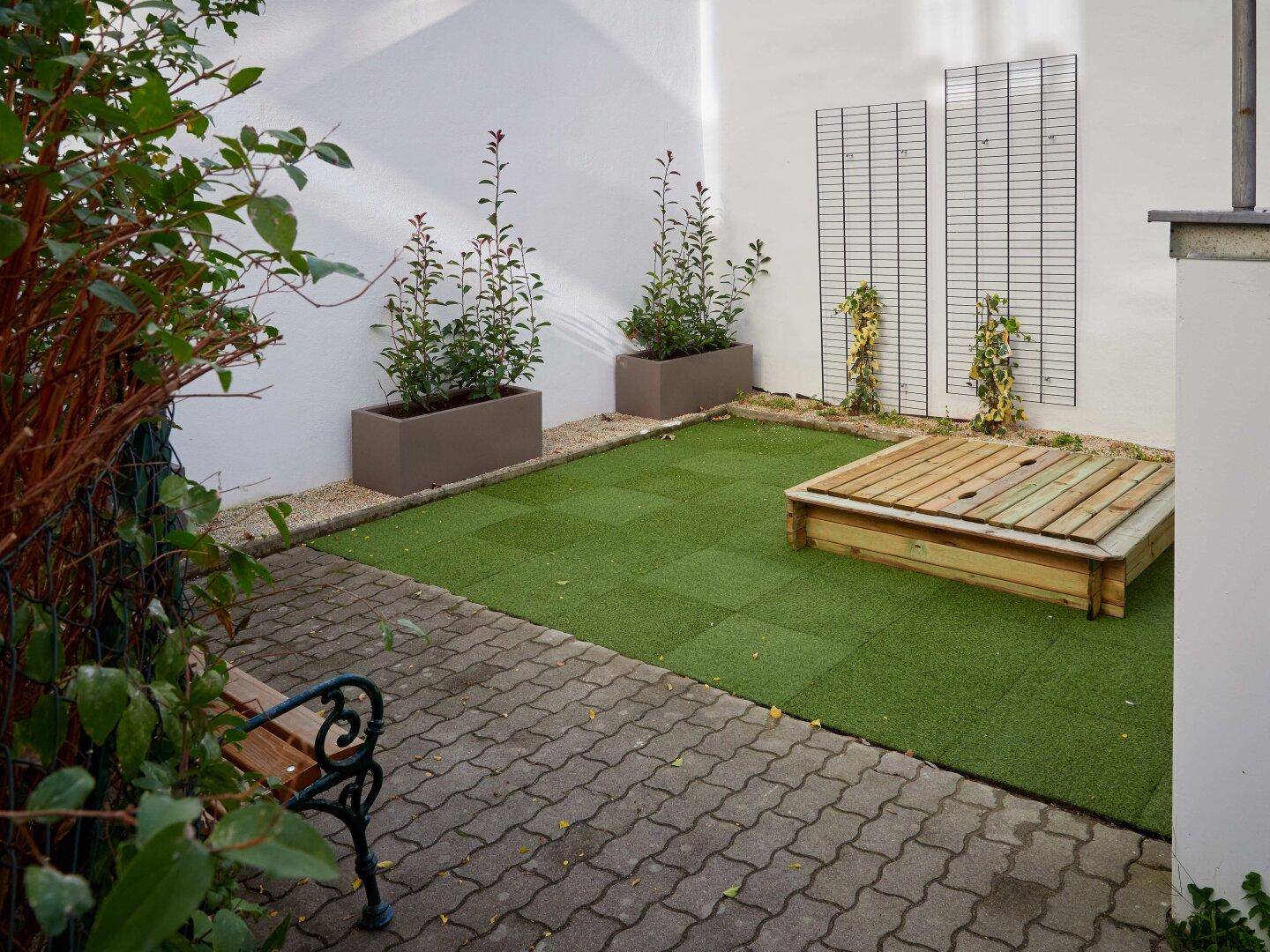 Kleiner Innenhofgarten mit Kunstrasen, Sandkasten und Pflanzen in Hochbeeten.
