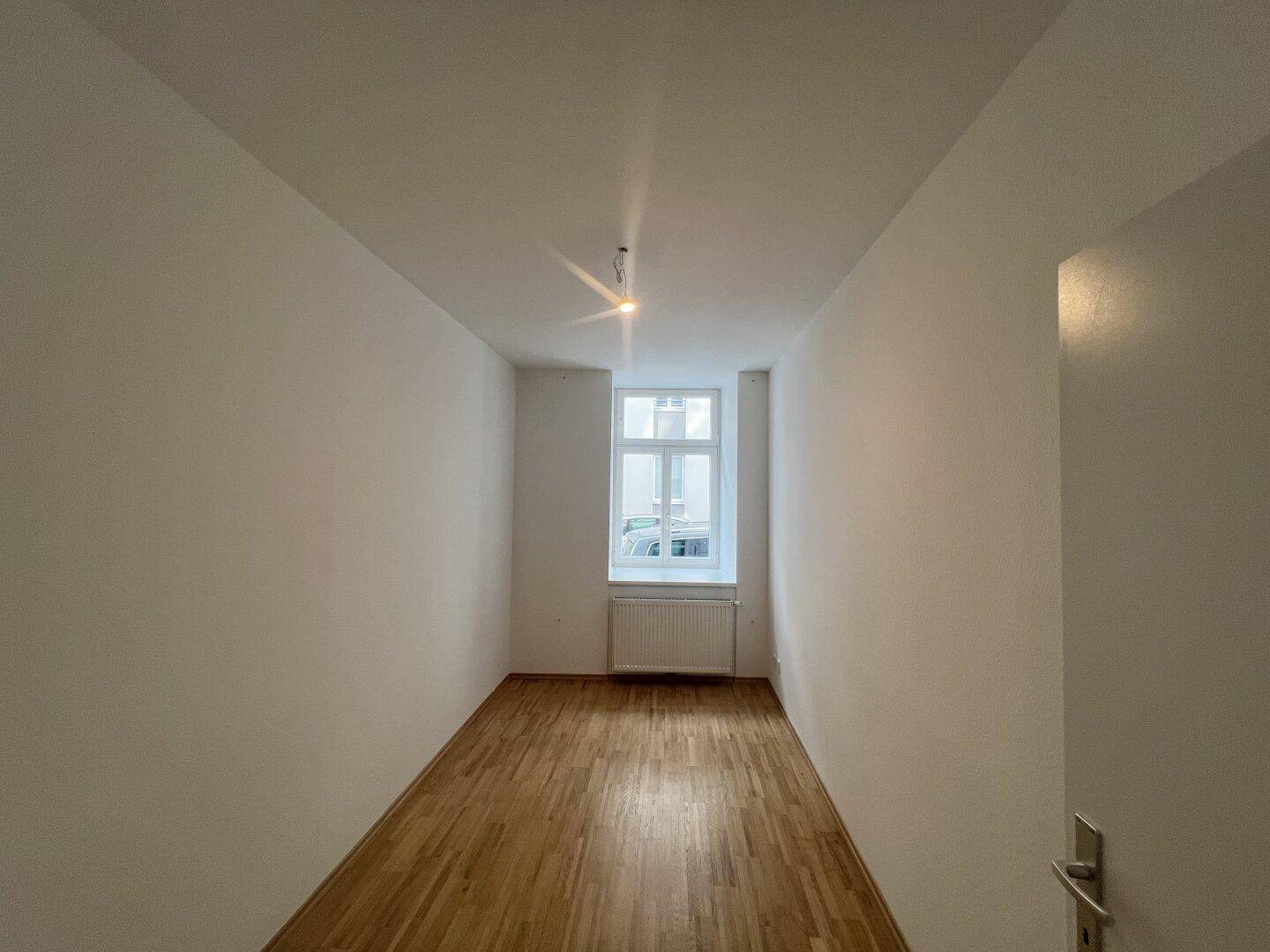 Schmaler Raum mit Holzboden und einem Fenster, ideal als Schlaf- oder Arbeitszimmer.