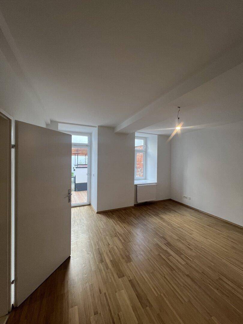 Geräumiger Raum mit Holzboden, weißen Wänden und Zugang zu einem Außenbereich.