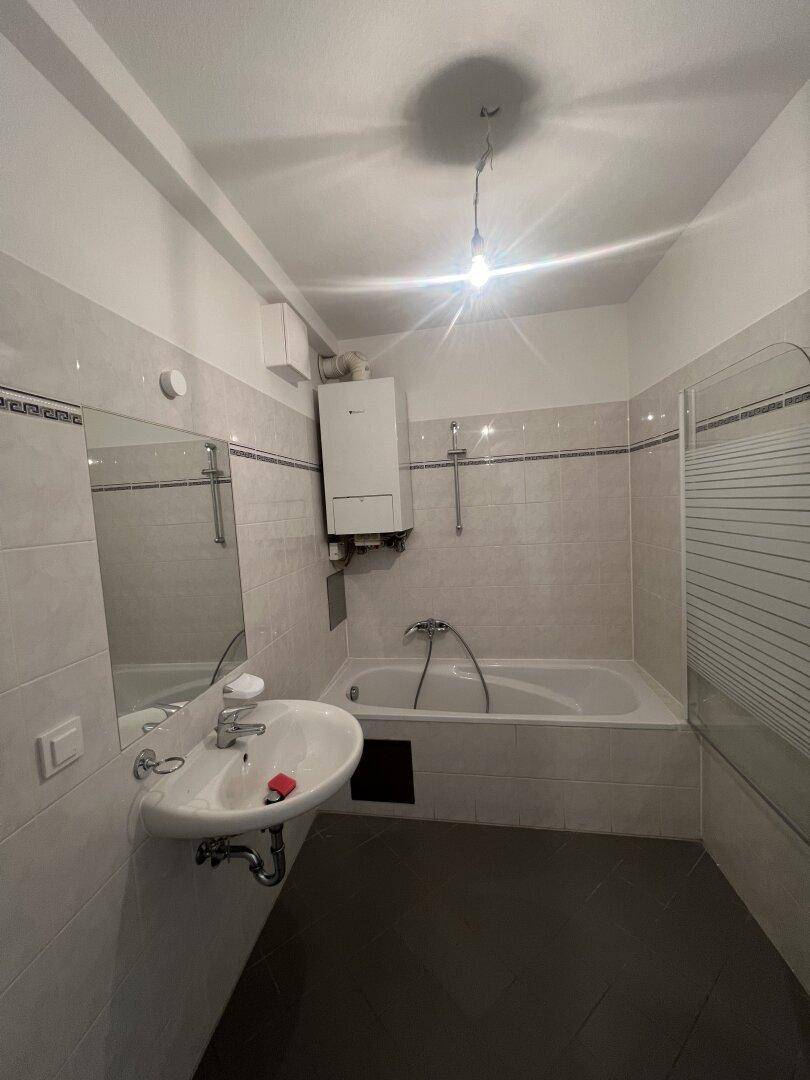 Badezimmer mit Badewanne, Waschbecken und hellen Fliesen, inklusive Warmwasserbereiter.