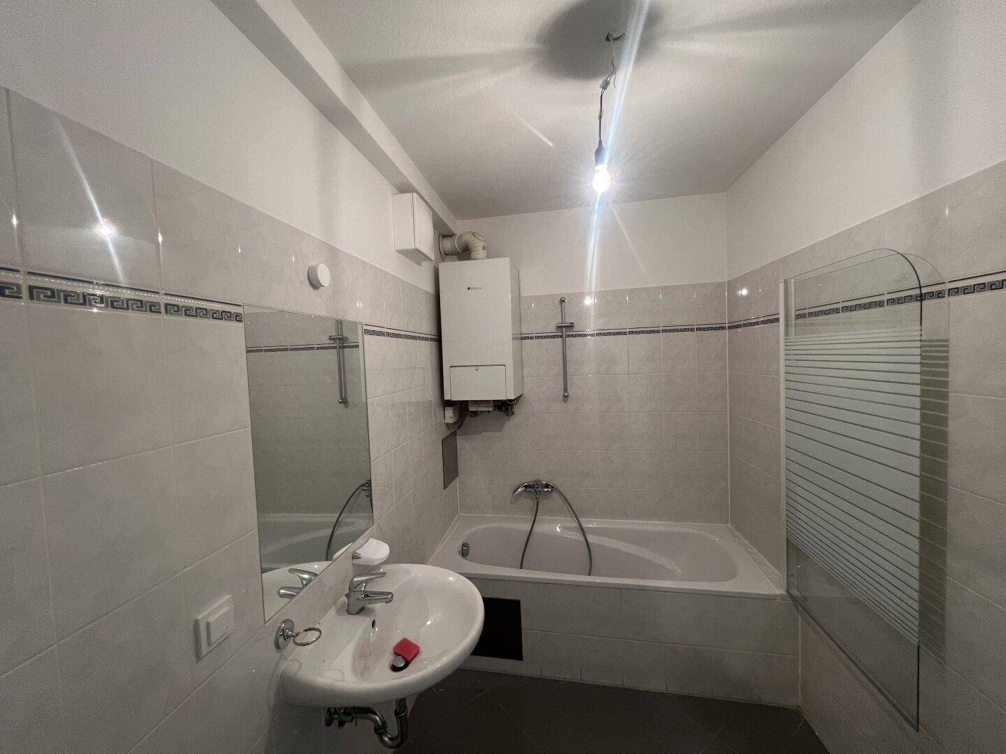 Badezimmer mit Badewanne, Waschbecken und hellen Fliesen, inklusive Warmwasserbereiter.