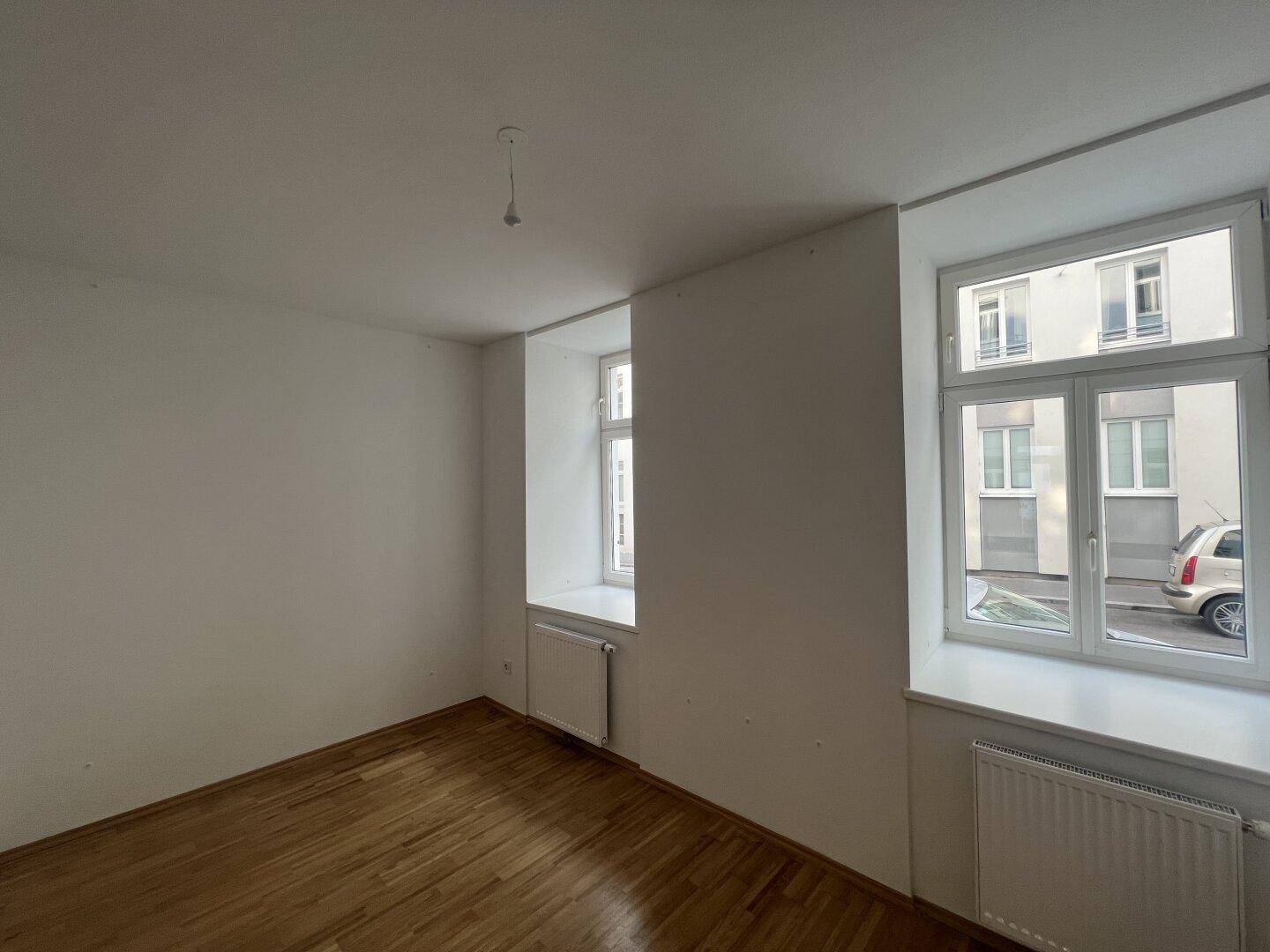 Helles Zimmer mit Holzboden und zwei Fenstern, die einen Blick auf die Umgebung bieten.