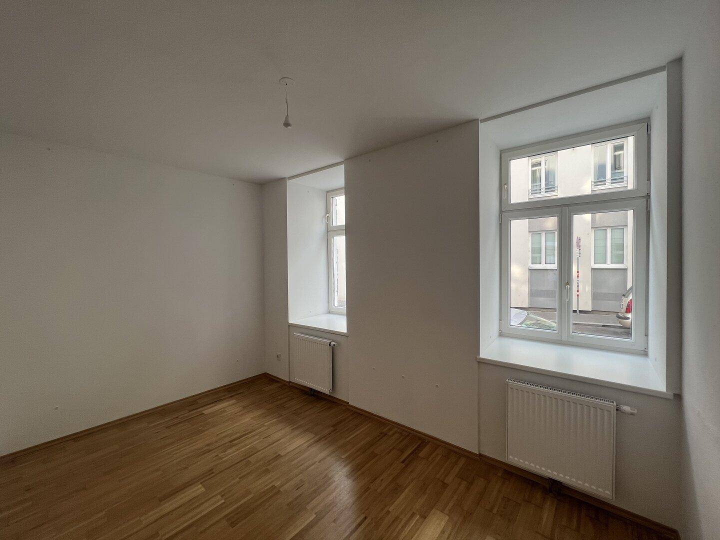 Helles Zimmer mit Holzboden und zwei Fenstern, die einen Blick auf die Umgebung bieten.