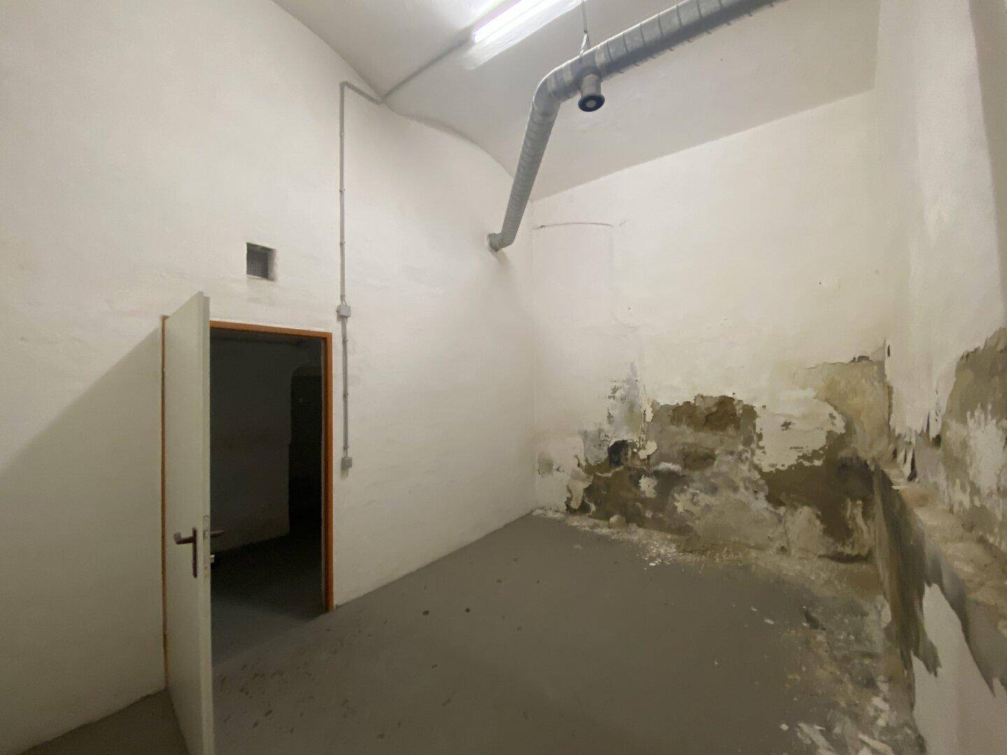 Kellerraum mit abblätternder Farbe an den Wänden, sichtbaren Lüftungsrohren und einer Tür.