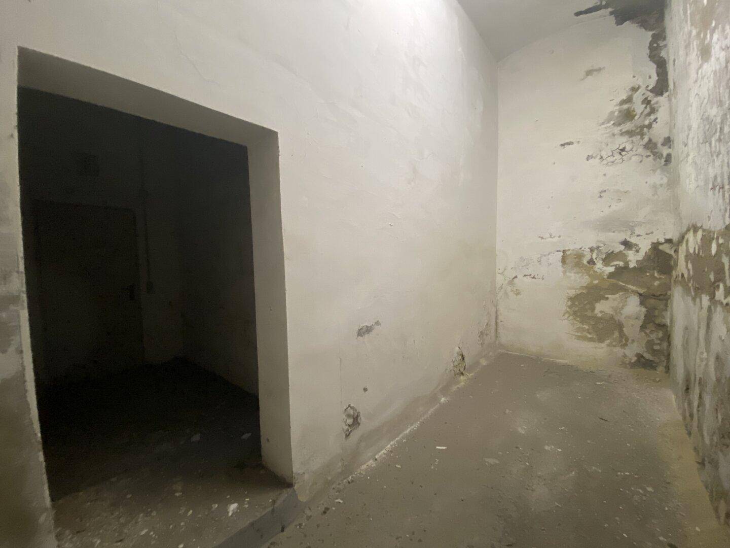 Schmaler Kellergang mit unverputzten Wänden und einem offenen Durchgang zu einem weiteren Raum.