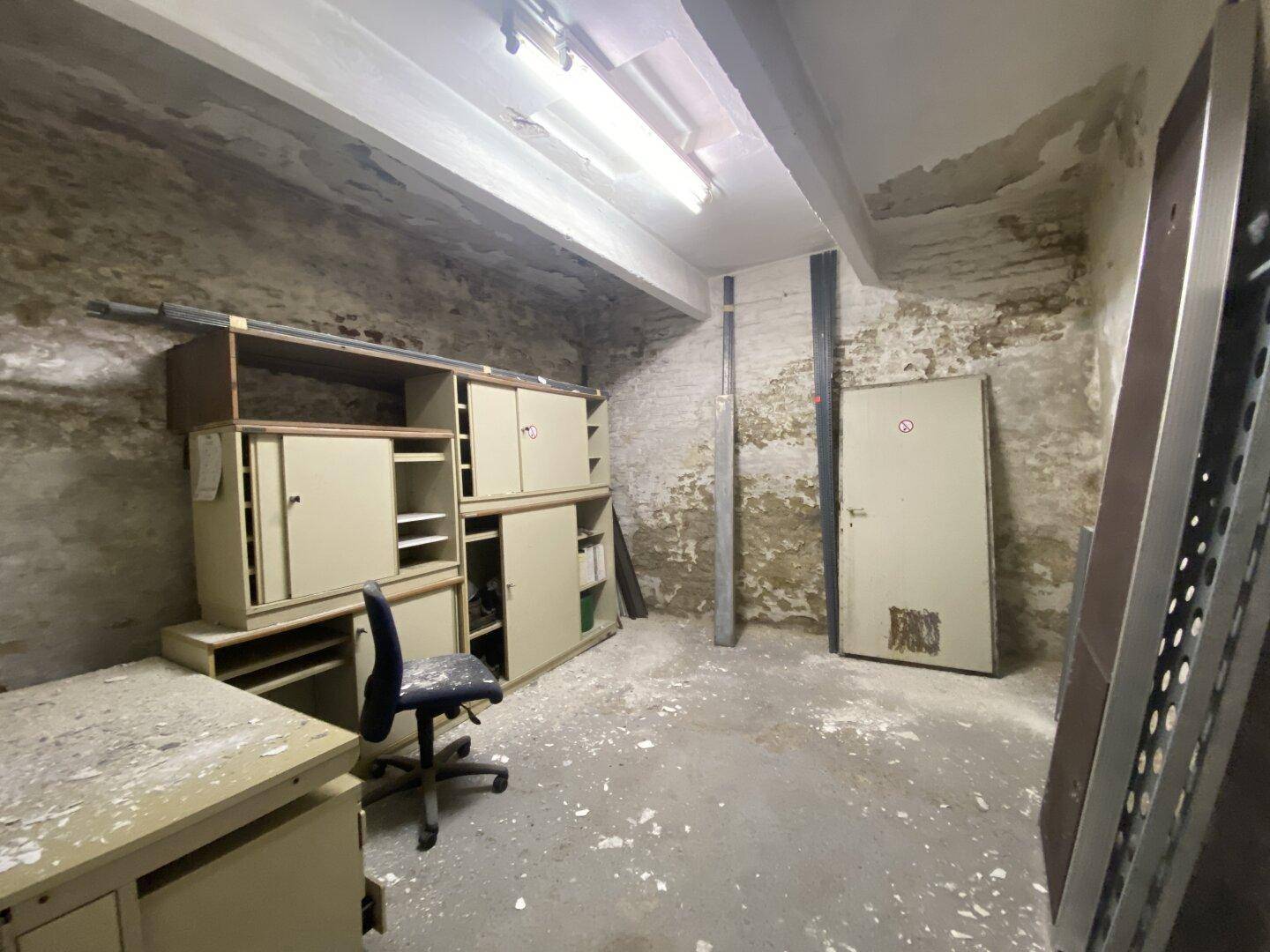 Kellerraum mit alten Büromöbeln, unverputzten Wänden und sichtbaren Deckenbalken.