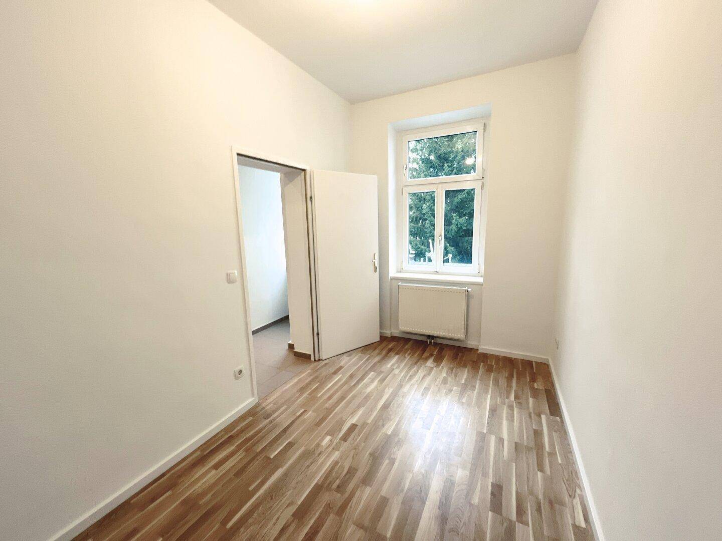 Helles Zimmer mit Parkettboden und Fenster zum grünen Außenbereich, ideal als Schlafzimmer.