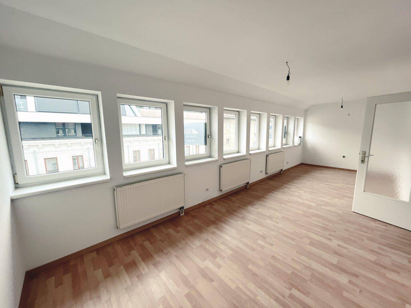 Geräumiges, helles Zimmer mit einer Reihe von Fenstern und hellem Laminatboden.