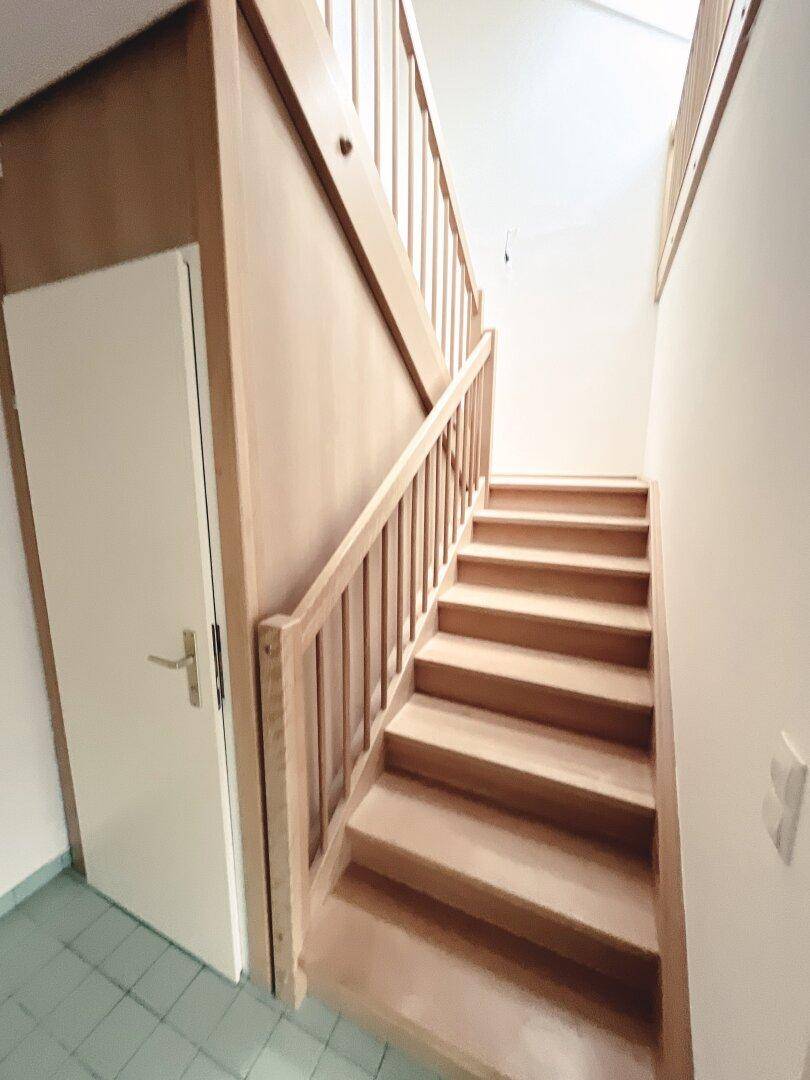Holztreppe führt in die obere Etage, mit hellem Holzgeländer und gefliestem Boden.