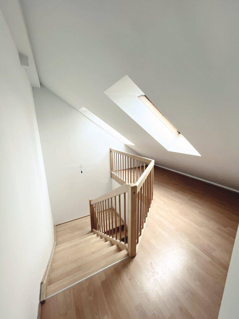 Helle Holztreppe führt zu einem Dachgeschossraum mit schrägen Wänden und Dachfenstern.
