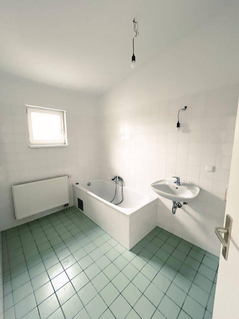 Badezimmer mit Badewanne, Waschbecken und Fenster, ausgestattet mit hellgrünen Fliesen.