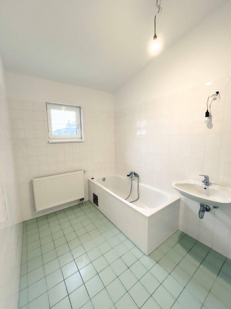 Einfaches Badezimmer mit Badewanne, Waschbecken und Fenster, mit hellgrünen Bodenfliesen.
