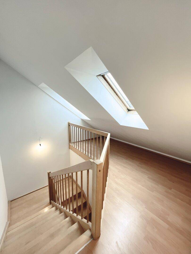 Holztreppe führt zu einem hellen Dachgeschossbereich mit schrägen Wänden und Dachfenstern.