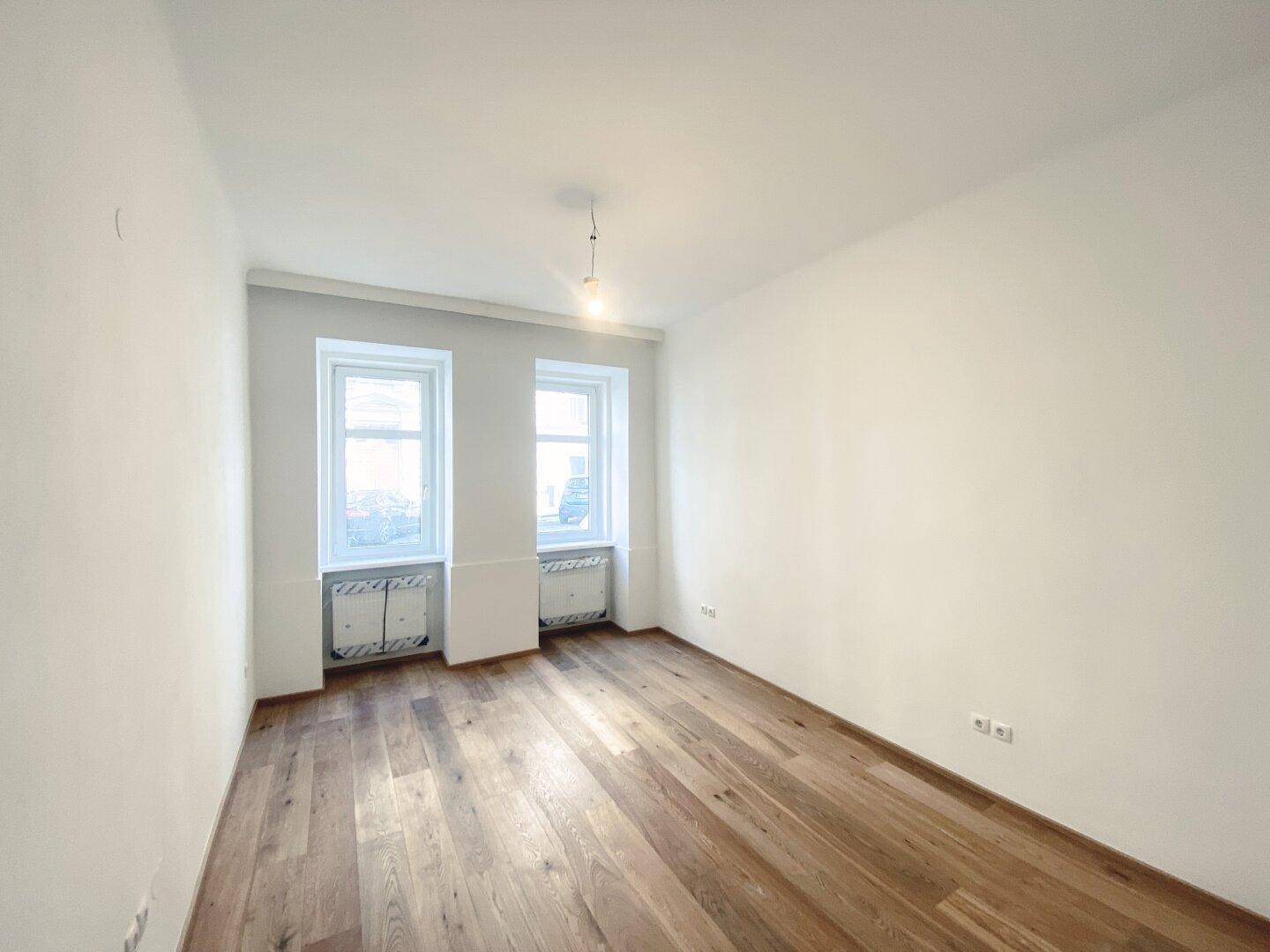 Geräumiges Zimmer mit Holzboden und zwei Fenstern, ideal für Wohn- oder Schlafbereich.