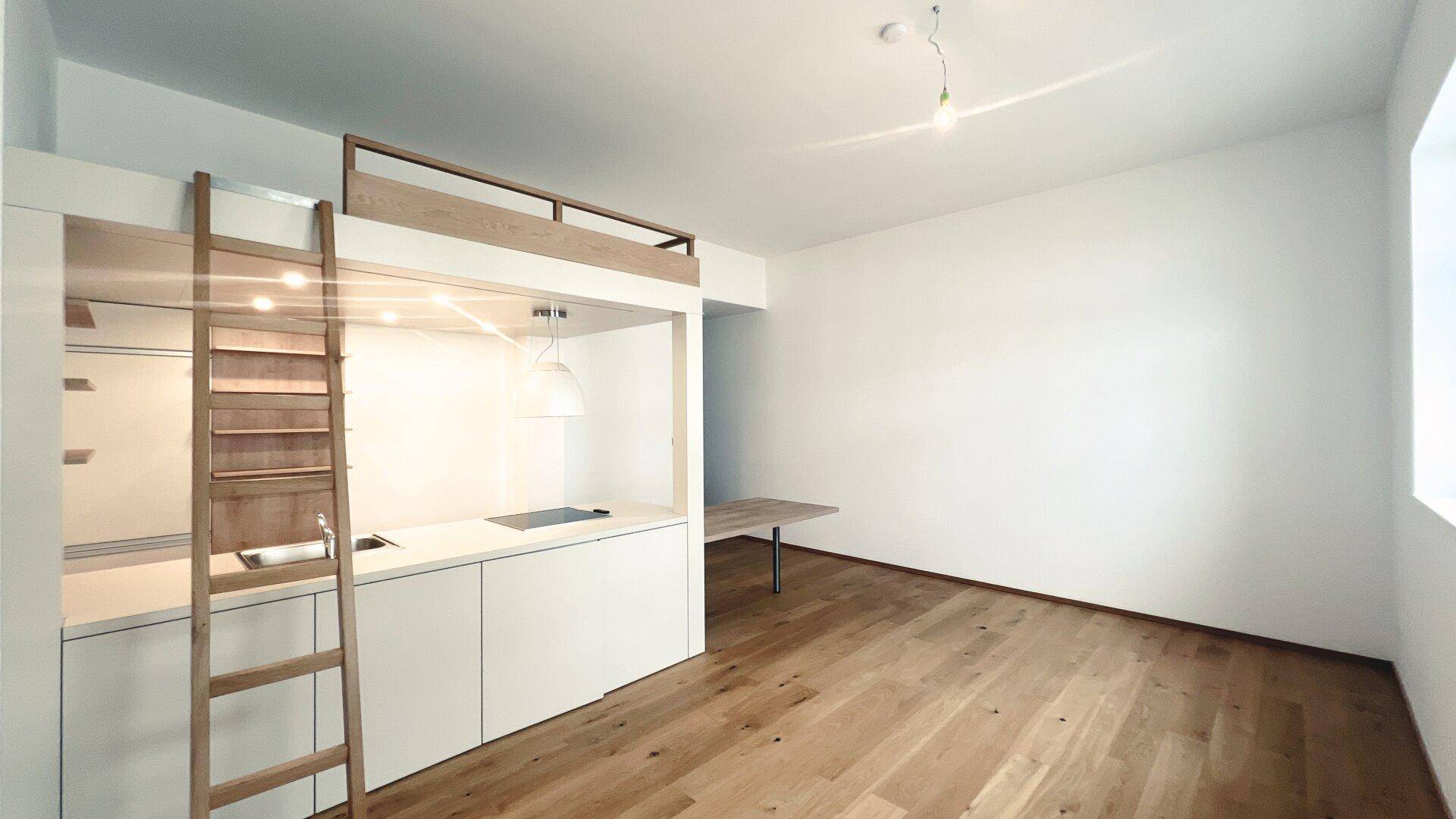 Heller Wohnbereich mit integrierter Küche und Hochbett, das über eine Holzleiter zugänglich ist.