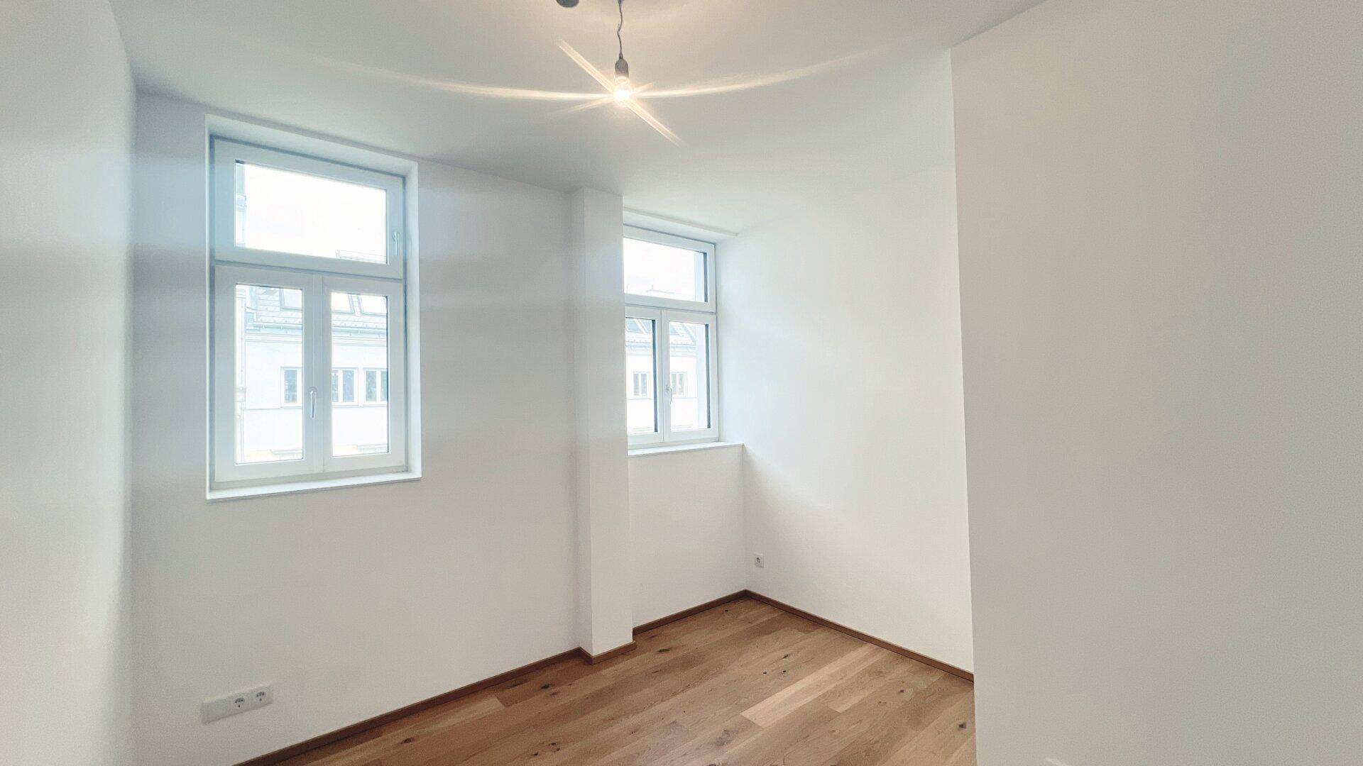 Lichtdurchflutetes Zimmer mit zwei Fenstern und Holzboden, bietet viel natürliches Licht.