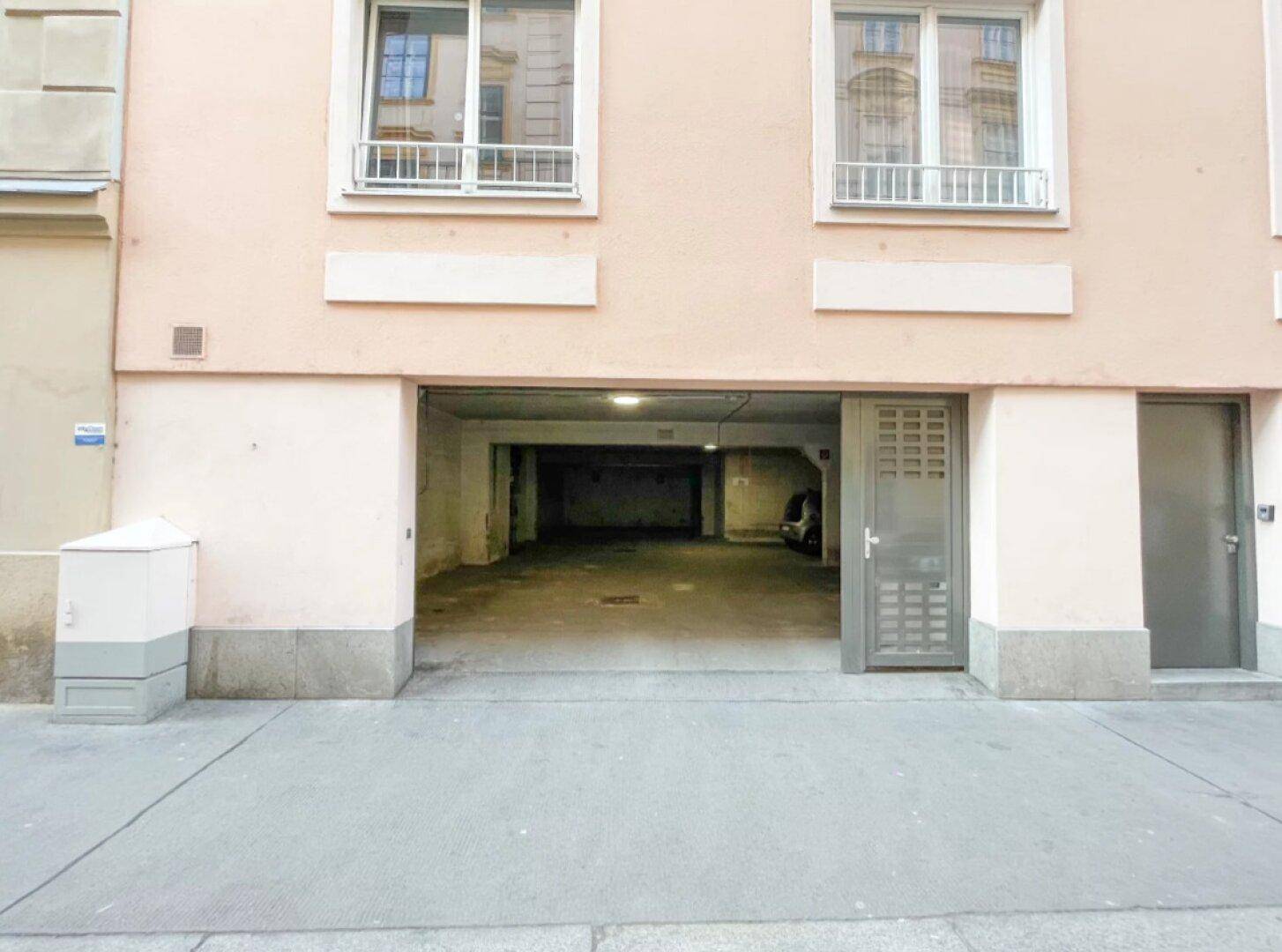 Gebäudefassade mit Garageneinfahrt auf Straßenniveau und Fenstern im Obergeschoss.