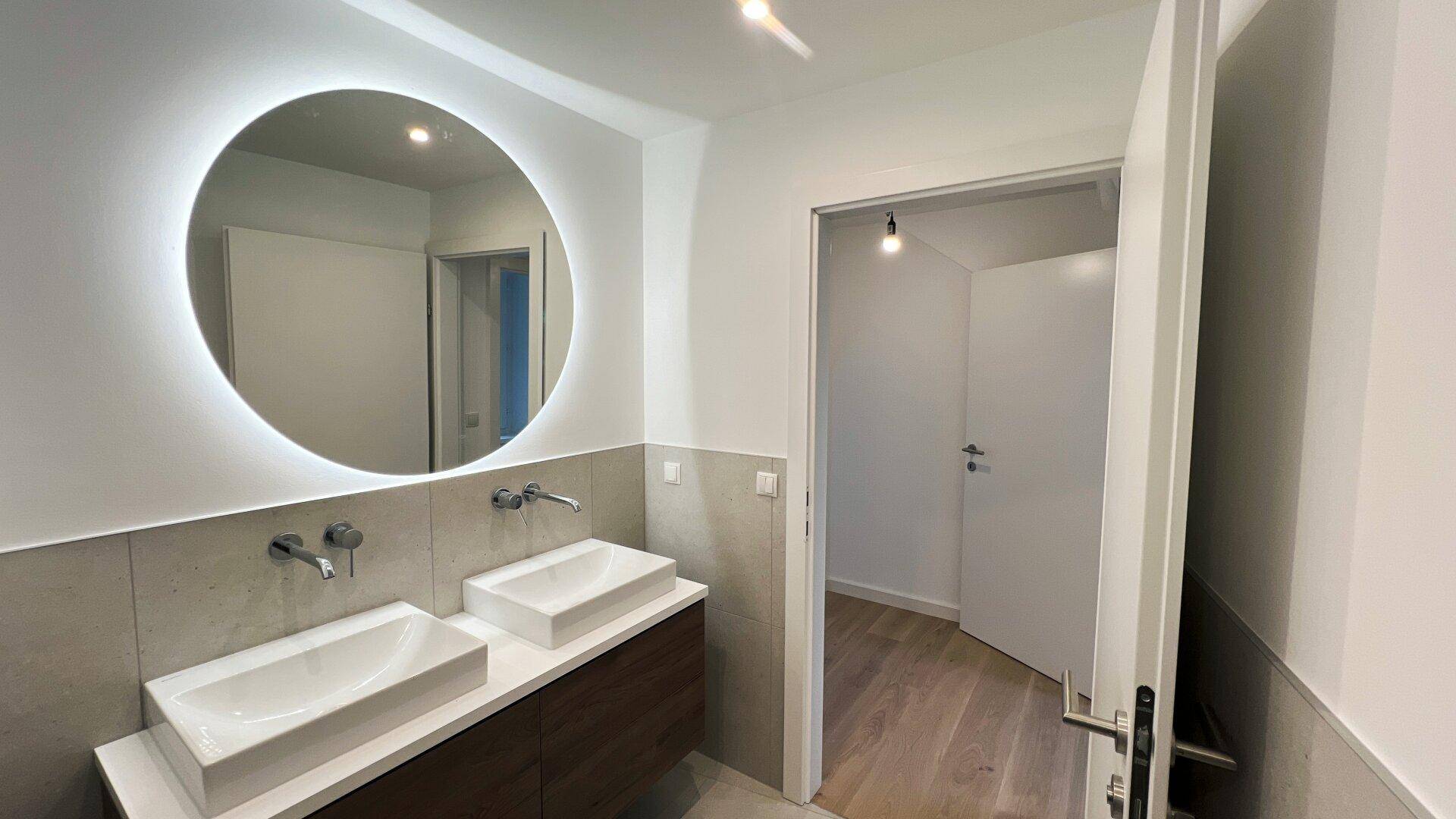 Modernes Badezimmer mit Doppelwaschbecken, beleuchtetem Spiegel und grauen Fliesen.