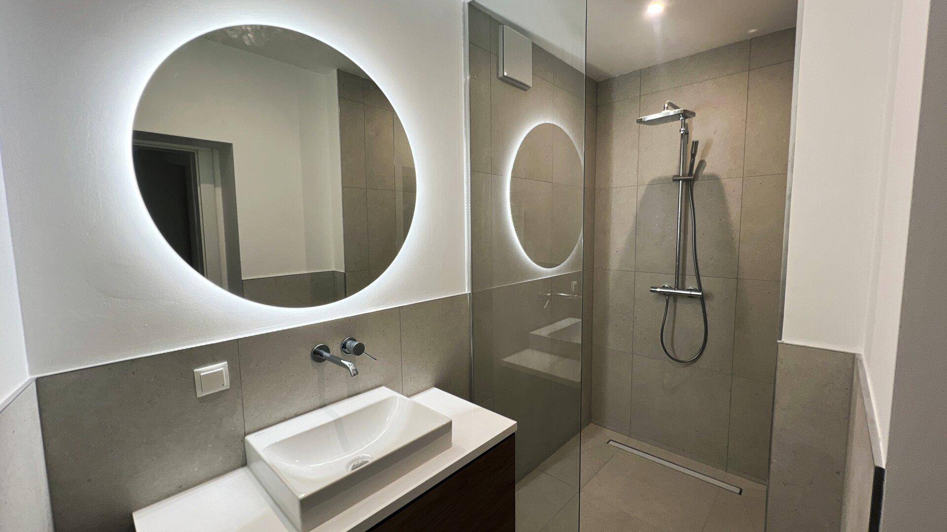 Modernes Badezimmer mit begehbarer Dusche, beleuchtetem Spiegel und grauen Fliesen.