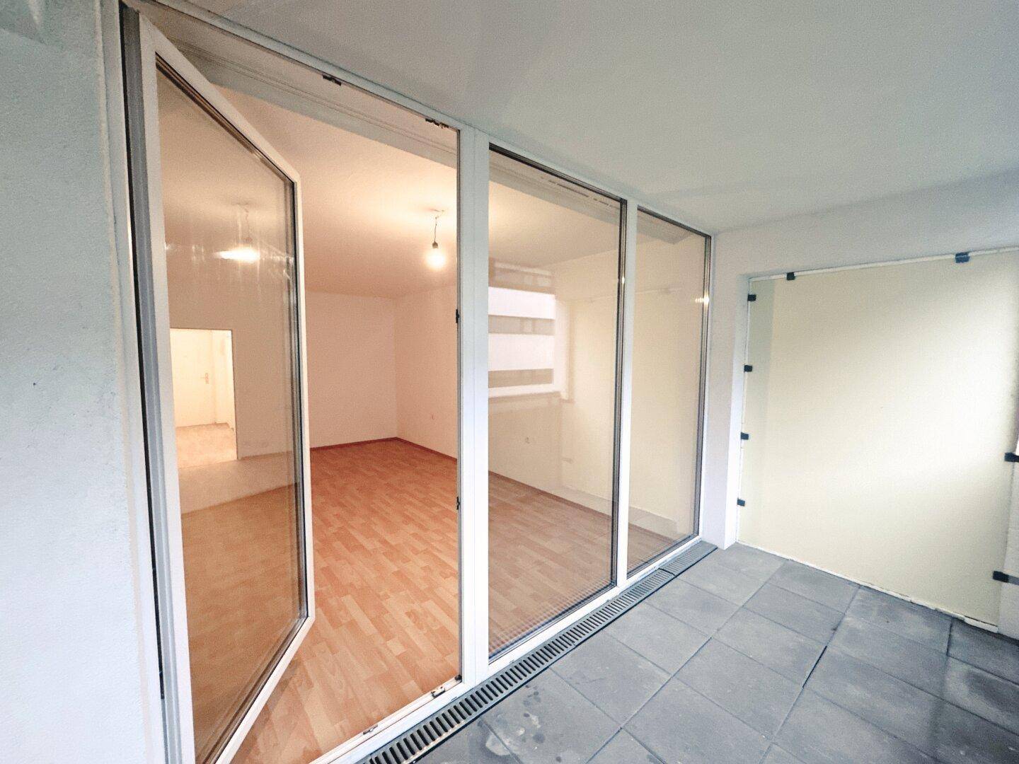 Balkon mit Fliesenboden und Blick in das helle, leere Zimmer mit Laminatboden.