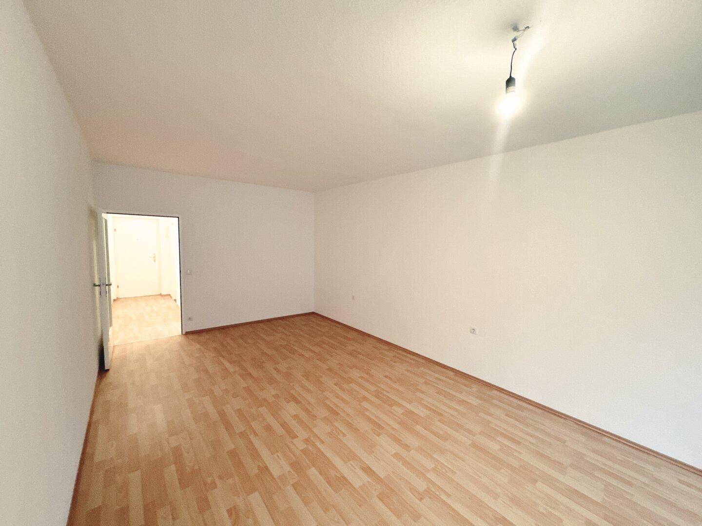 Geräumiges, leeres Zimmer mit hellem Laminatboden und einem offenen Durchgang zu einem weiteren Bereich.