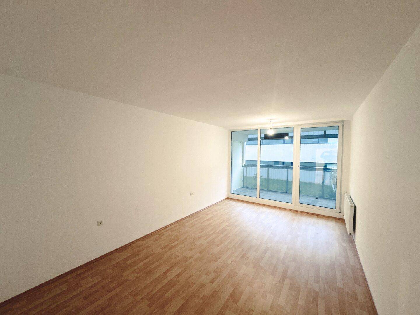 Helles, leeres Wohnzimmer mit Laminatboden und großem Fenster mit Balkonzugang.