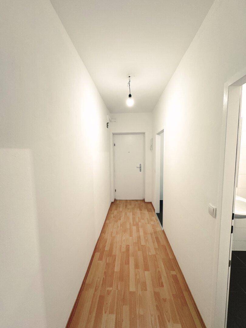 Langer, heller Flur mit Laminatboden und weiß gestrichenen Wänden, führt zur Eingangstür.