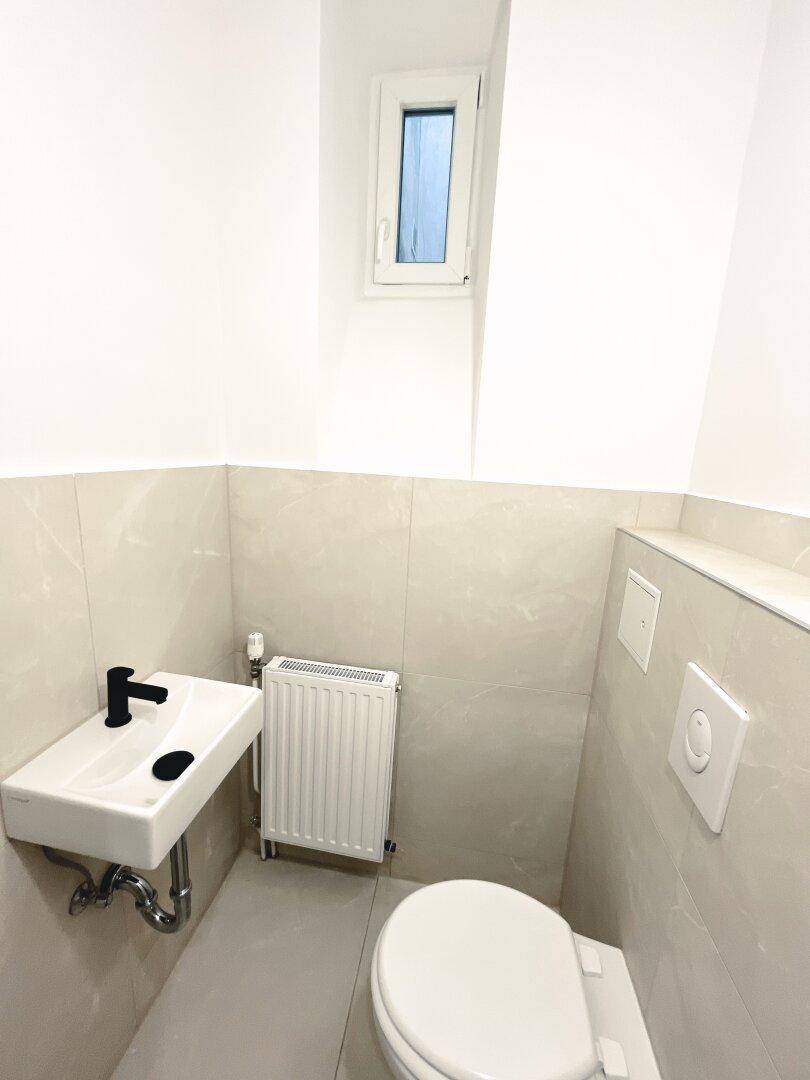 Modernes Gäste-WC mit hellem Fliesenboden, wandhängendem WC und kleinem Waschbecken.