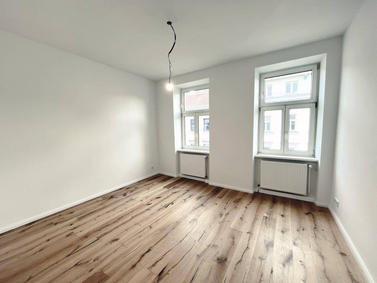 Helles Zimmer mit zwei Fenstern, Holzboden und weiß gestrichenen Wänden, in gutem Zustand.