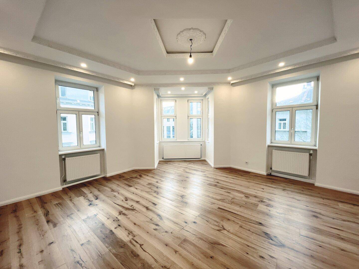 Geräumiges Wohnzimmer mit Parkettboden, dekorativer Decke und mehreren Fenstern für viel Licht.