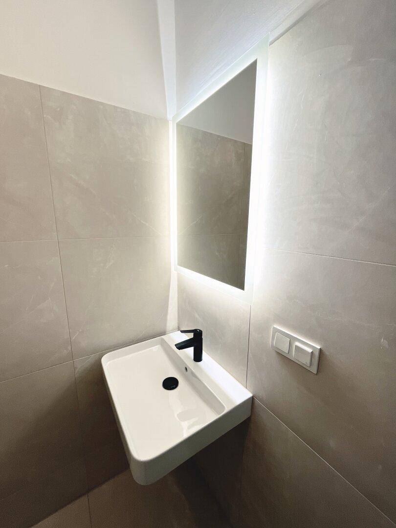 Modernes Badezimmer mit beleuchtetem Spiegel, Waschbecken und hellen Fliesen.