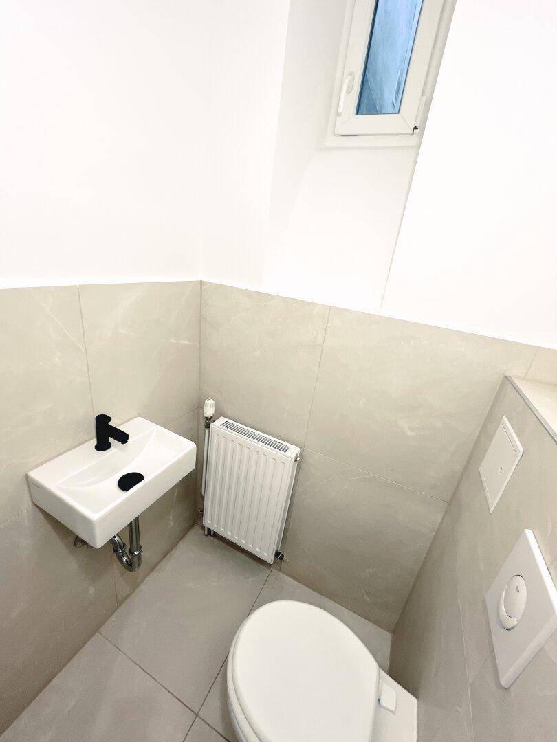 Modernes Gäste-WC mit hellem Fliesenboden, wandhängendem WC und kleinem Waschbecken.