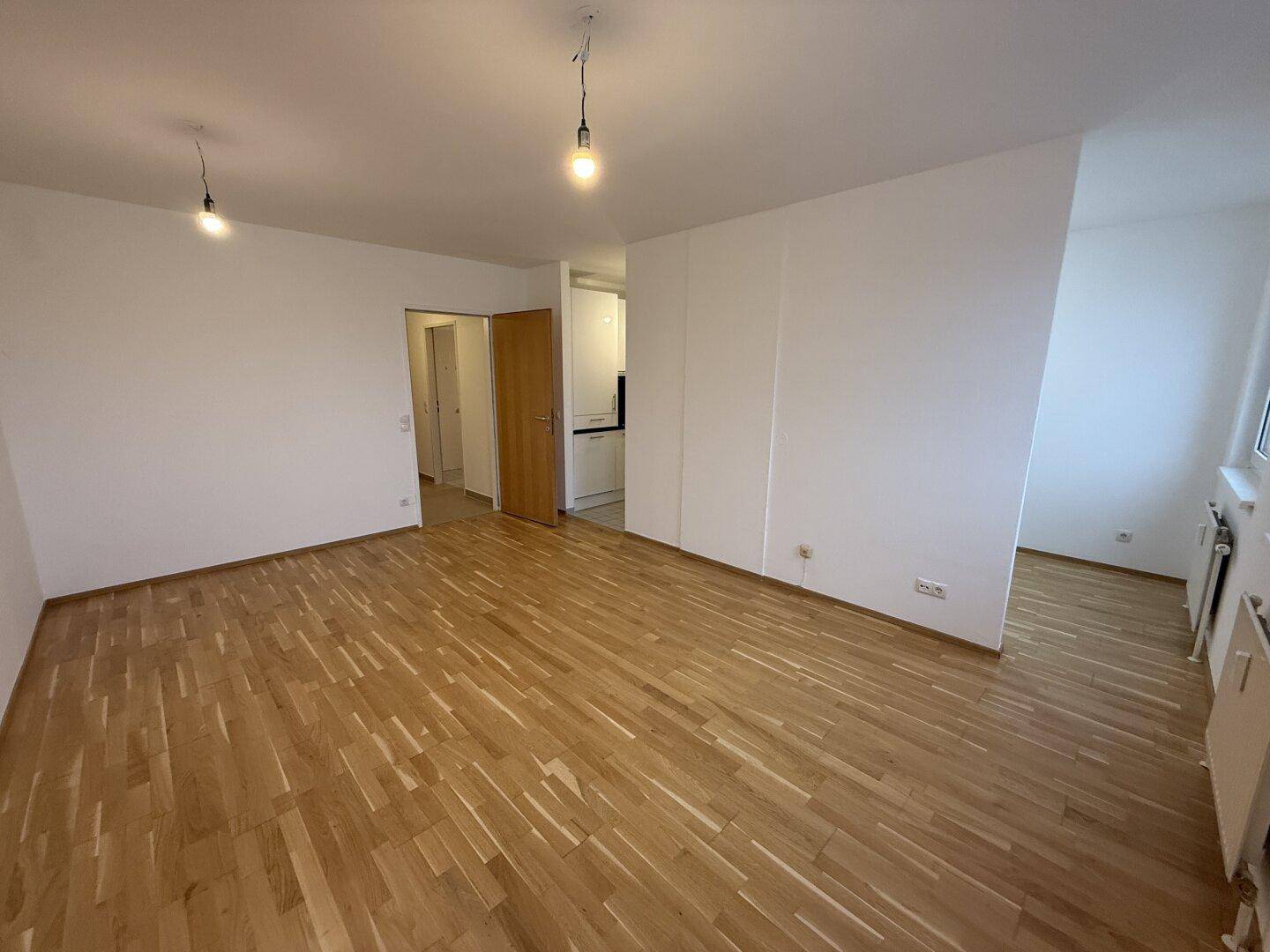 Helles, leeres Wohnzimmer mit hellem Parkettboden und weiß gestrichenen Wänden.