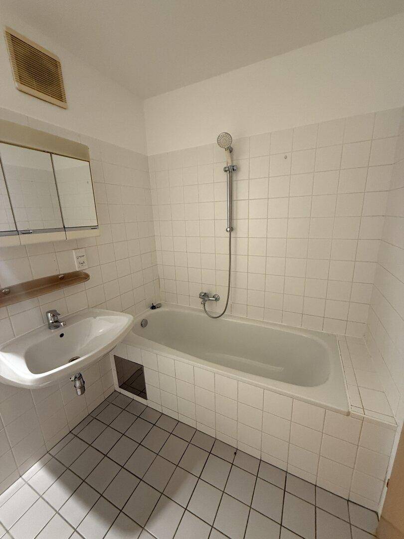Helles Badezimmer mit weißen Fliesen, Badewanne mit Duschfunktion und Waschbecken.