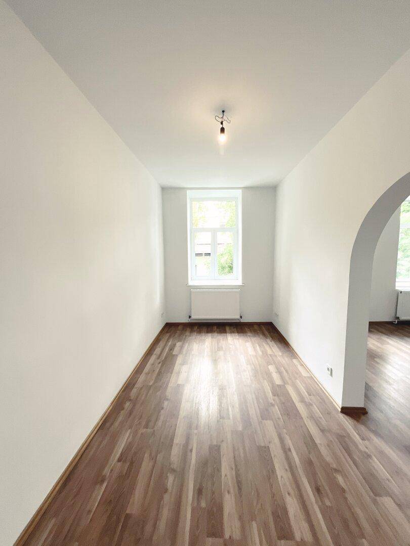Heller Raum mit Laminatboden und einem Fenster, verbunden durch einen eleganten Bogen zu einem weiteren Bereich.