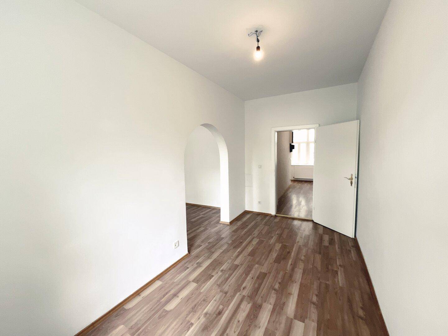 Lichtdurchfluteter Raum mit Laminatboden, einem Bogen und einer offenen Tür zu einem angrenzenden Bereich.