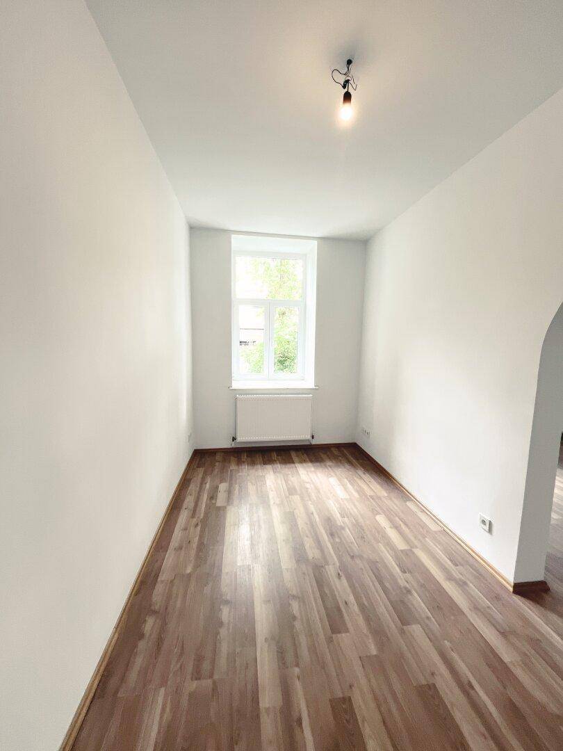 Heller Raum mit Laminatboden und einem Fenster, verbunden durch einen eleganten Bogen zu einem weiteren Bereich.