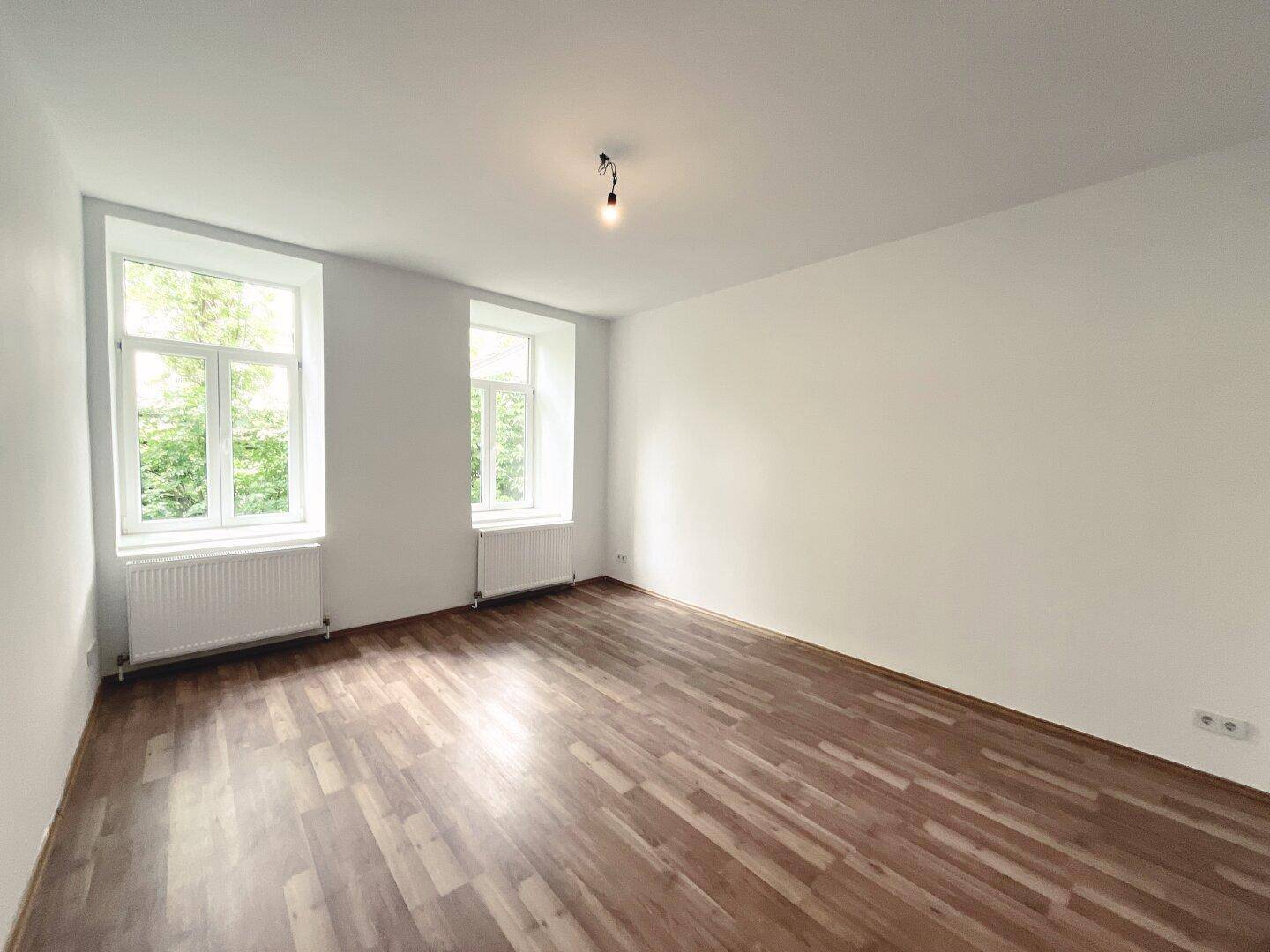 Helles Zimmer mit Laminatboden und zwei großen Fenstern, ideal als Wohn- oder Schlafzimmer.