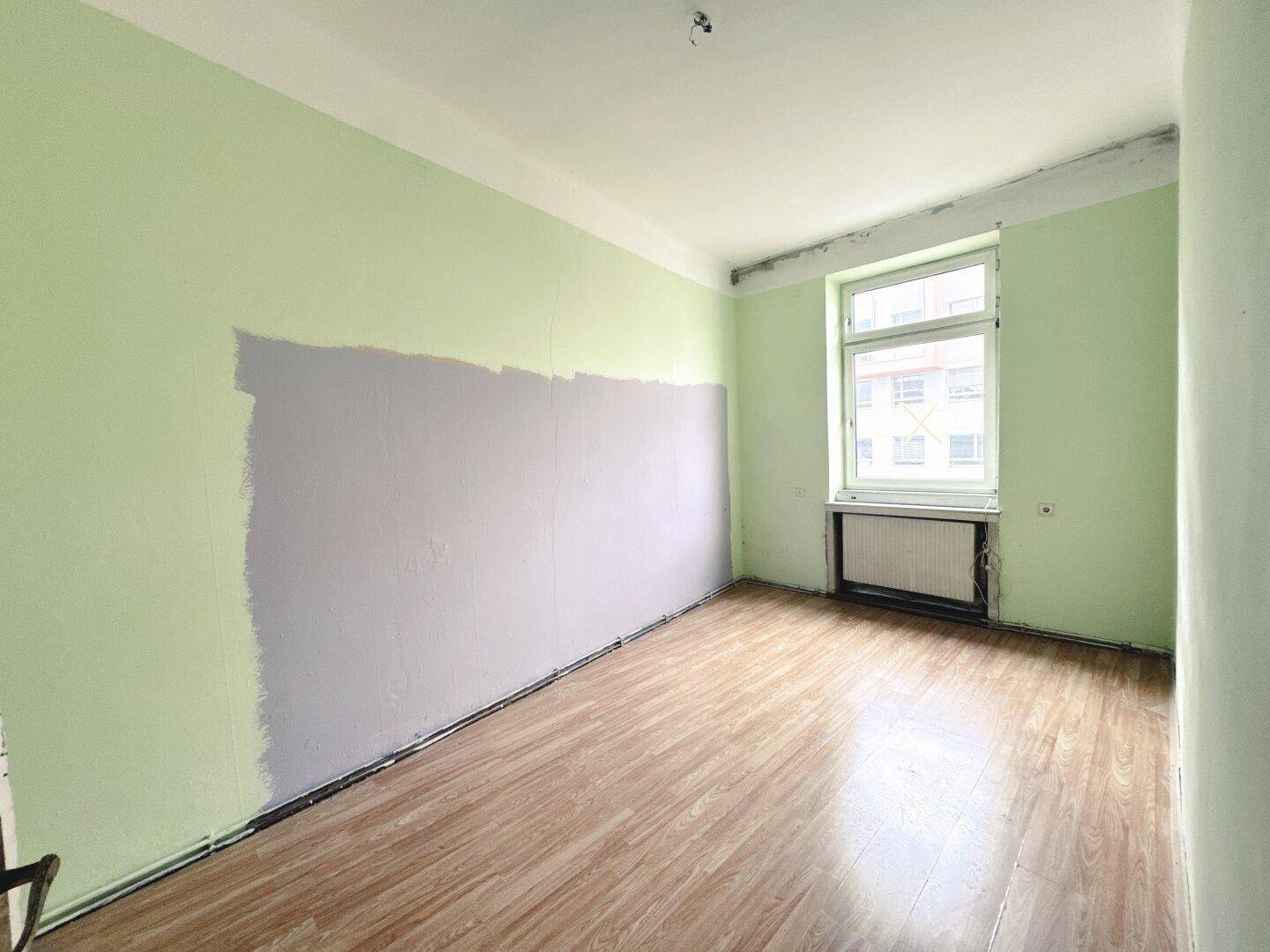 Helles Zimmer mit grünen Wänden und Laminatboden, teilweise grau gestrichen, mit Fenster.