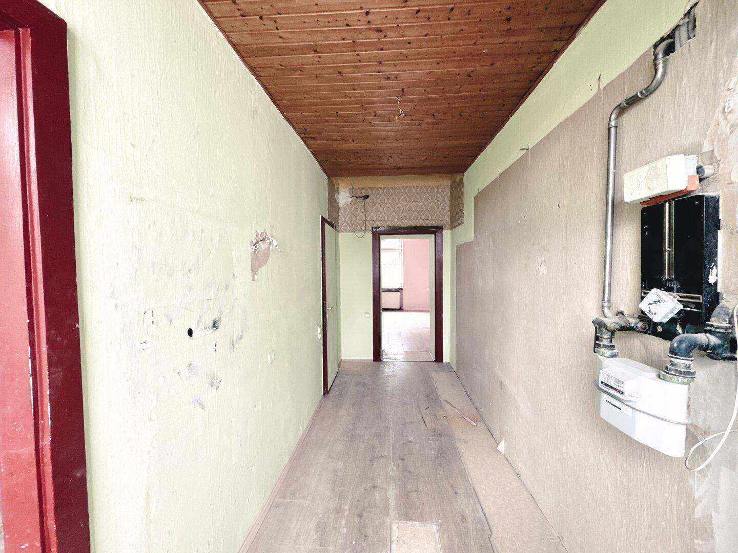 Flur mit Holzdecke und Dielenboden, zeigt Gaszähler und Leitungen, renovierungsbedürftig.