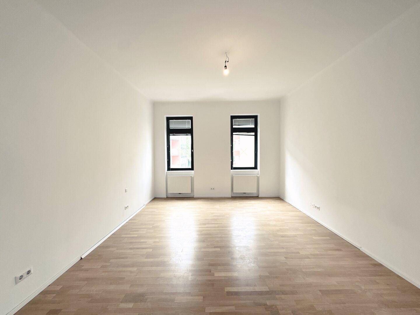Heller, leerer Raum mit Parkettboden und zwei Fenstern, bietet viel Tageslicht.