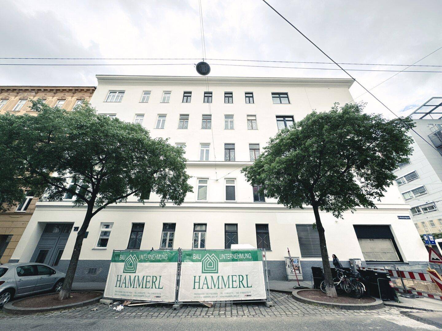 Fassade eines Wohngebäudes mit Bäumen davor und Bauzäunen im Vordergrund.