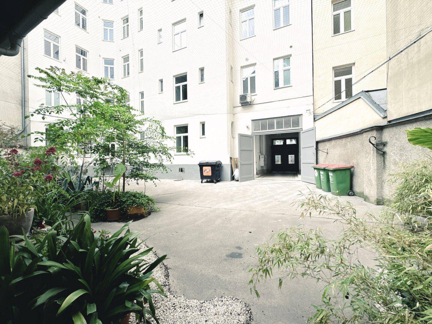 Innenhof eines Wohngebäudes mit Pflanzen und Mülltonnen, umgeben von Fassaden.