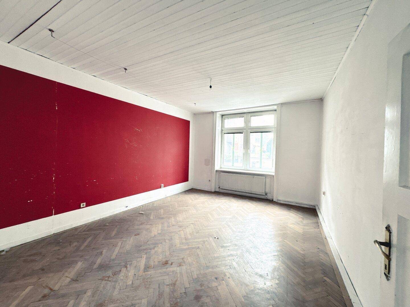 Leeres Zimmer mit Fischgrätparkett, roter Akzentwand und weiß gestrichener Holzdecke.