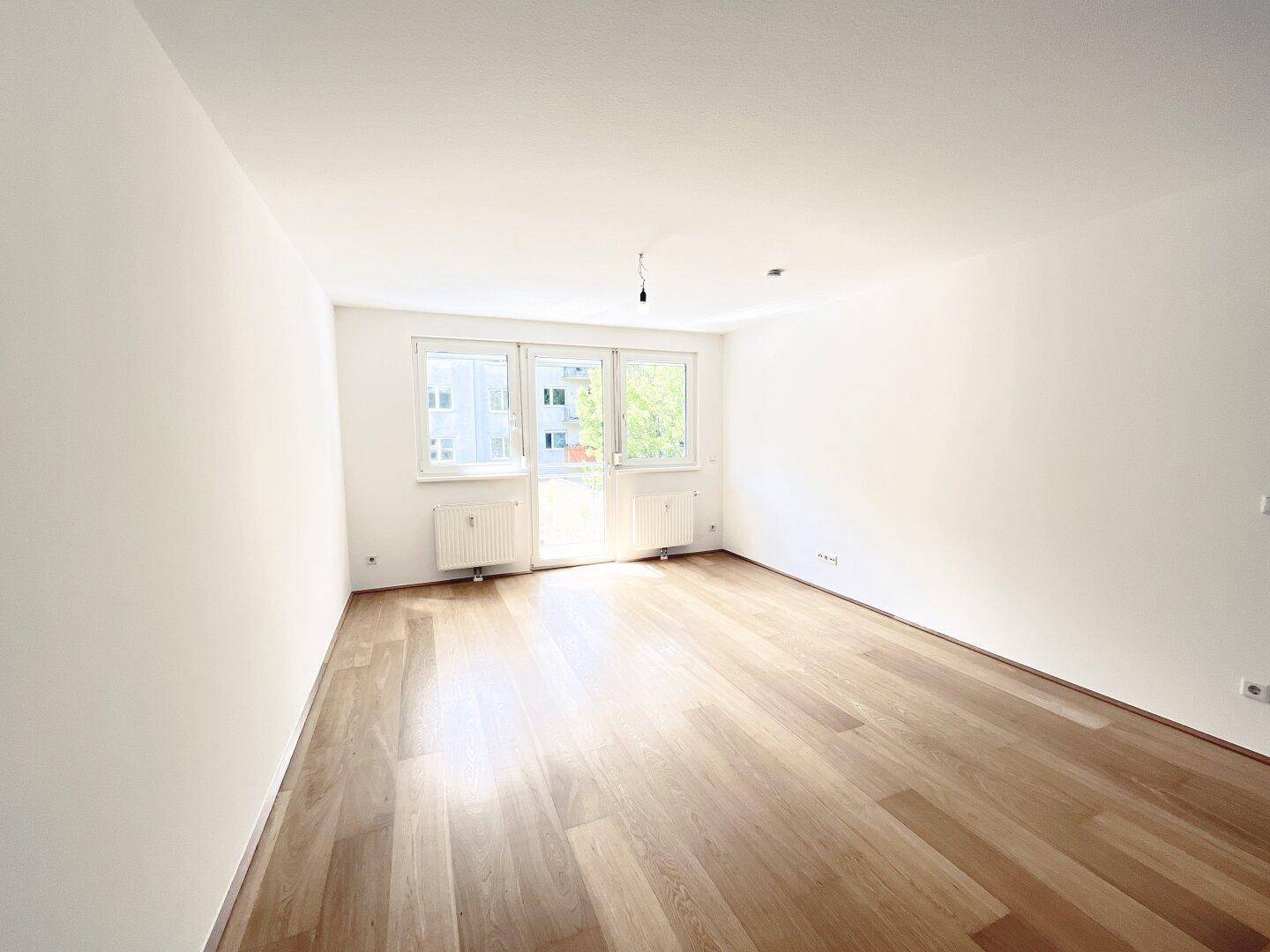 Helles, leeres Zimmer mit Parkettboden und großen Fenstern, die viel Tageslicht hereinlassen.