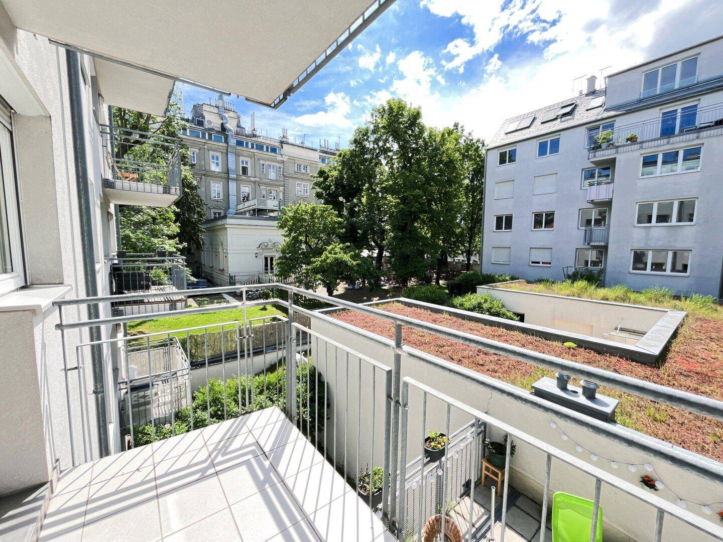 Heller Balkon mit Fliesenboden und Blick auf grüne Dächer und umliegende Gebäude.