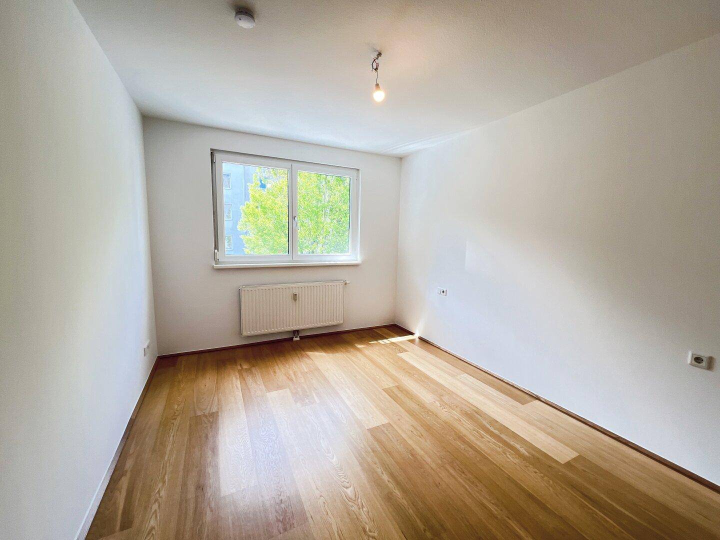 Helles Schlafzimmer mit Parkettboden und Fenster mit Blick ins Grüne.