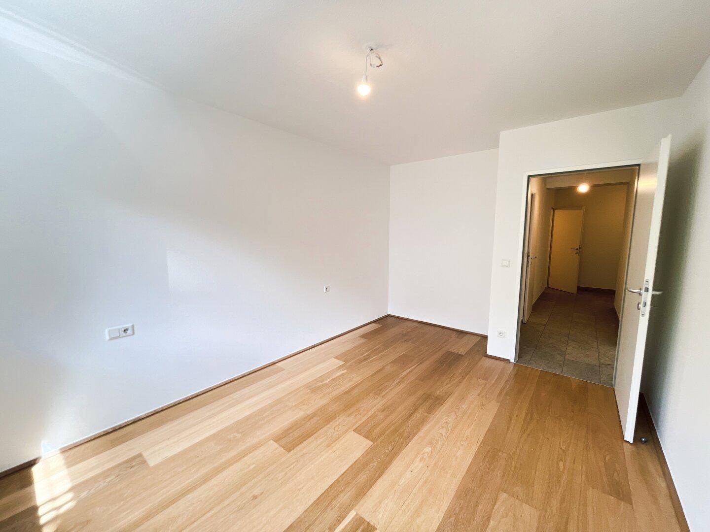 Helles Zimmer mit Holzboden und offener Tür, die in den Eingangsbereich führt.