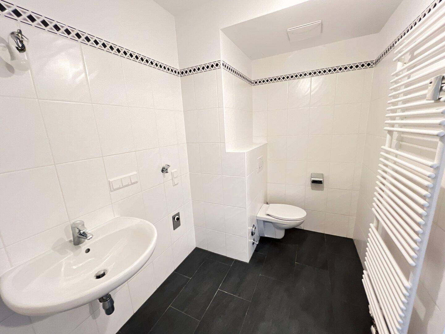 Badezimmer mit WC, Waschbecken, beheiztem Handtuchhalter und dunklem Fliesenboden.